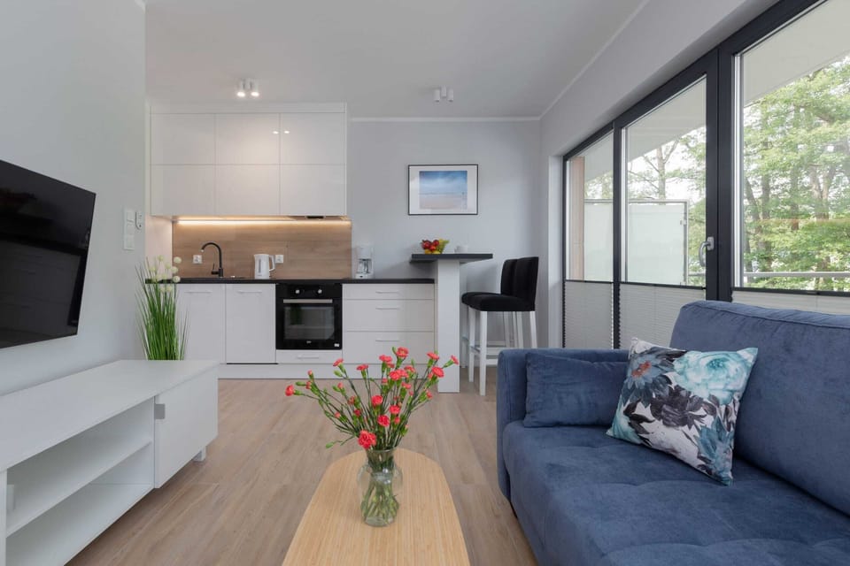Another cozy living room contains a gray sofa, a wooden coffee table with flowers, a mounted TV, and large windows letting in sunlight.