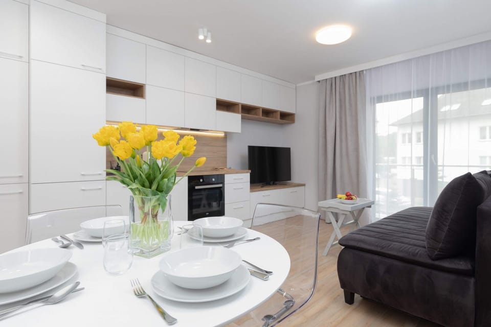 Close-up of a dining table with elegant white tableware, in the distance you can see a kitchenette, a TV and sofa.
