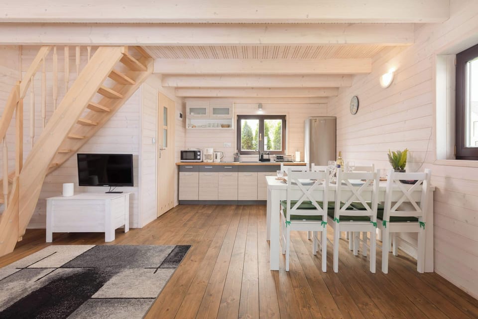 A spacious open-plan living area with a cozy seating zone, a dining table, and a fully equipped kitchenette. Light wooden walls and ceilings create a warm, Scandinavian-style atmosphere. Large windows bring in natural light.