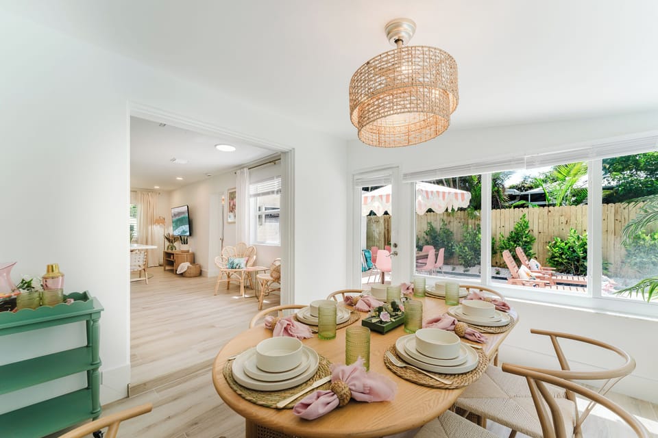 Designer dining room with seating for six, framed by oversized picture windows overlooking lush tropical landscaping and the pool. Thoughtfully styled with a statement woven chandelier, vibrant tropical artwork, and a charming cocktail cart.