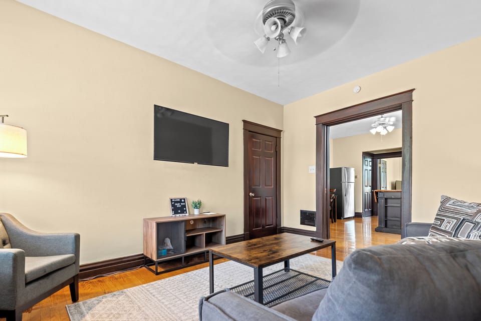 This reverse living-room angle shows the wall-mounted TV, gray sofa and armchair, wood coffee table, pale area rug, hardwood floors, and open doorway into the kitchen.