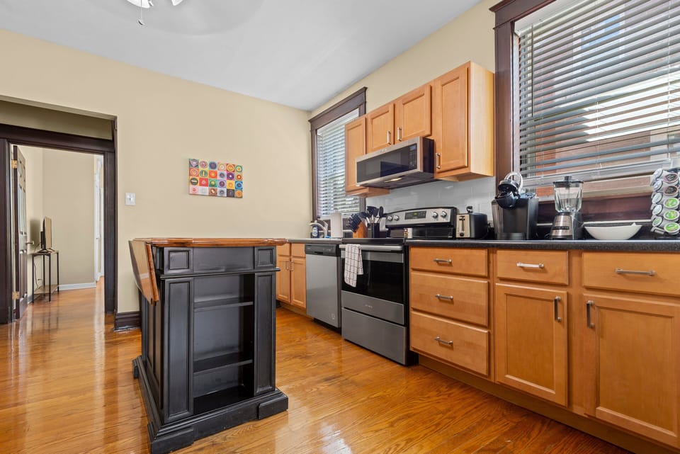 The kitchen features light wood cabinetry, dark counters, stainless range, microwave and dishwasher, coffee maker with pod storage, blender, double sink, and freestanding island.