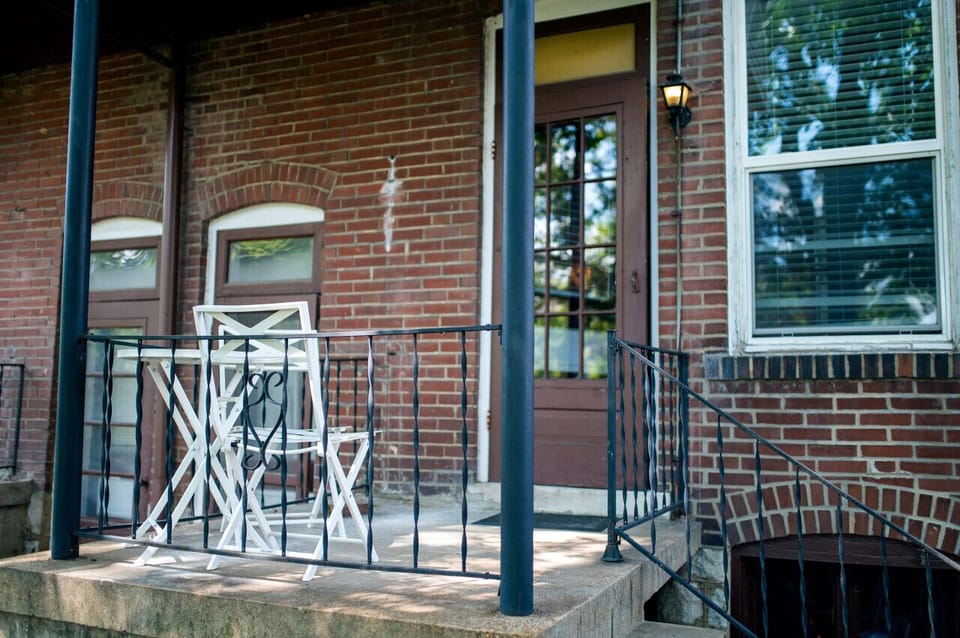 The covered concrete porch provides a white bistro table and chairs beside the windowed entrance, brick wall, and black metal railing.