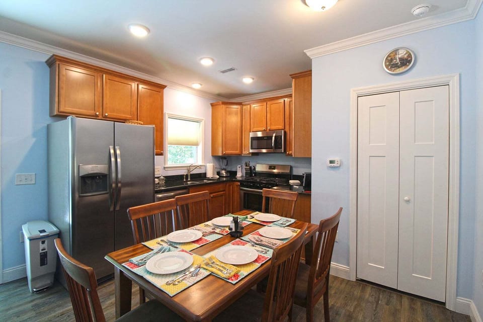 Dining table set for four beside the fully equipped kitchen with granite countertops, stainless refrigerator, and wood cabinets. Open layout connects the main living areas.