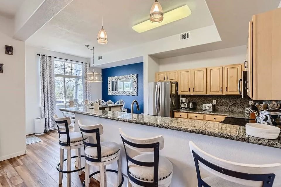Modern kitchen and dining area with a bar-height granite countertop, four barstools, stainless steel appliances, light wood cabinets, and a blue accent wall.