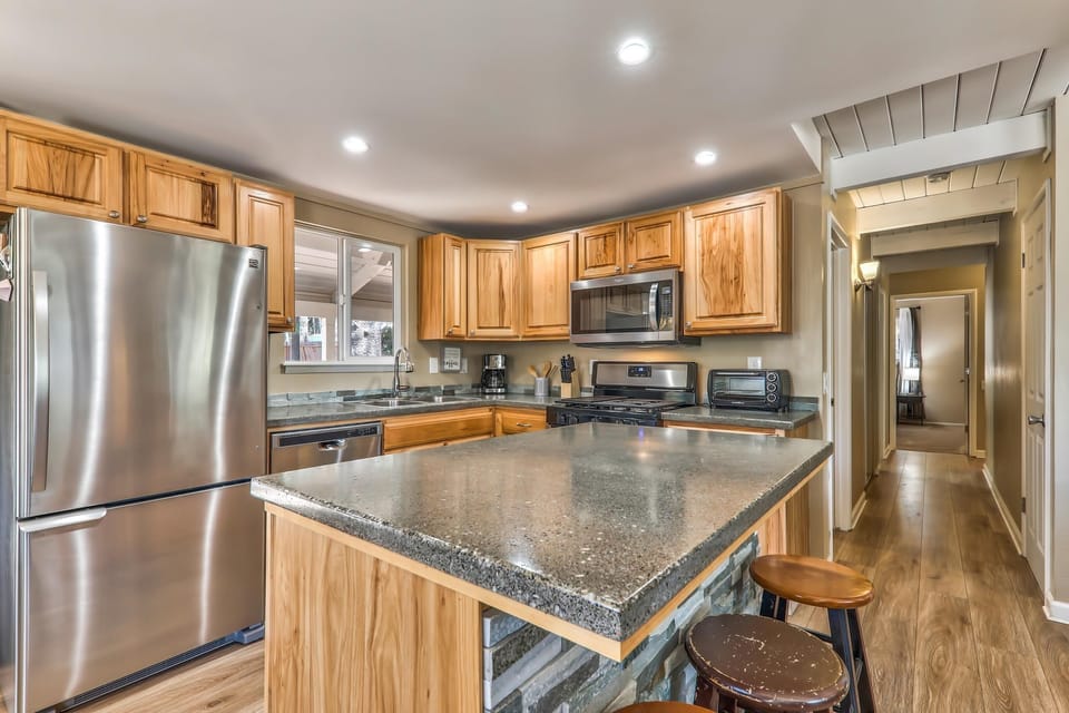Concrete countertop island with wood-paneled base and three wooden stools in kitchen with stainless appliances.