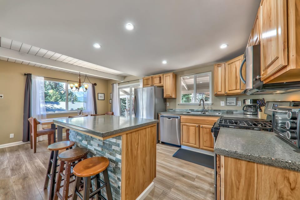 Kitchen features natural wood cabinets, stone-textured island with three wooden stools, and stainless steel appliances.