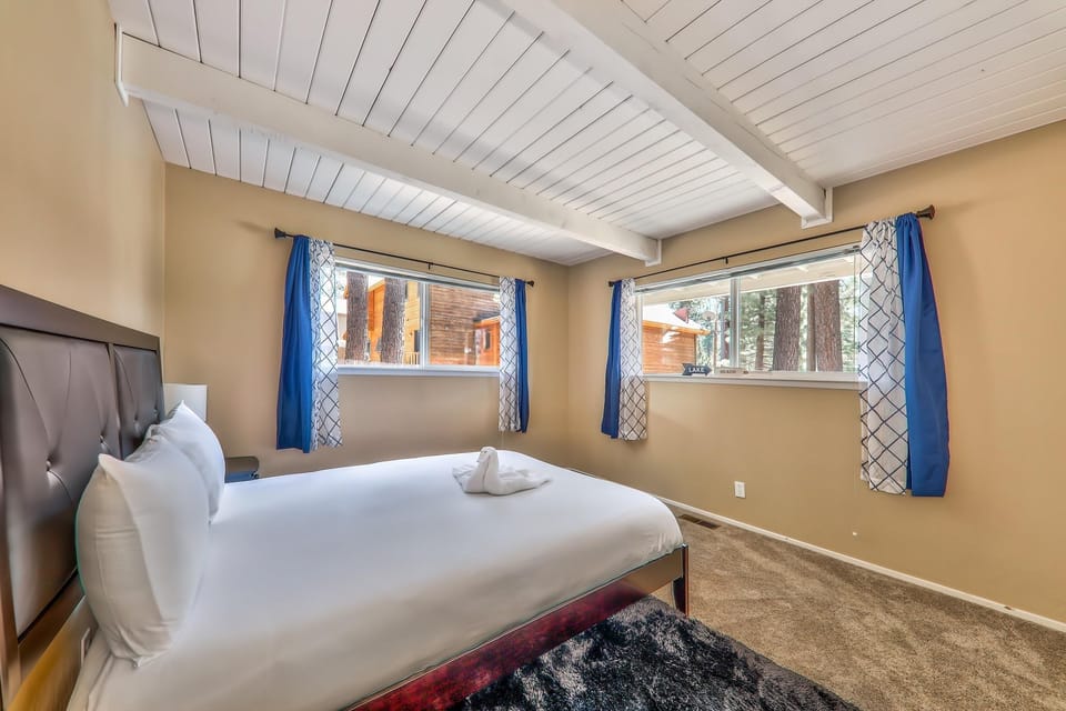 Natural light fills the bedroom with beige walls, white plank ceiling, blue and white patterned curtains, and a dark leather bed frame.