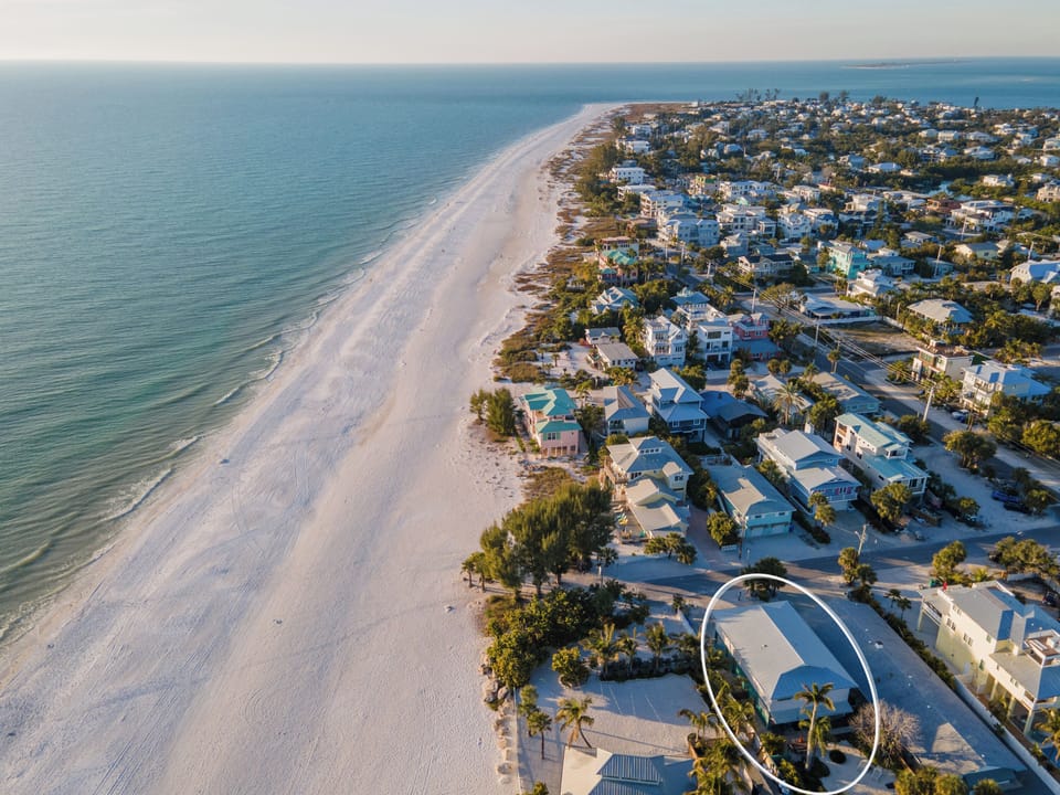 Stunning aerial view captures pristine white sand beach meeting turquoise waters, with charming coastal homes nestled among palm trees.