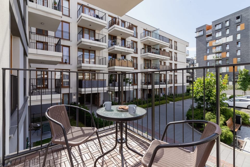 A balcony with a small table and chairs, surrounded by a black metal railing, offering a view of modern apartment buildings and a green courtyard.