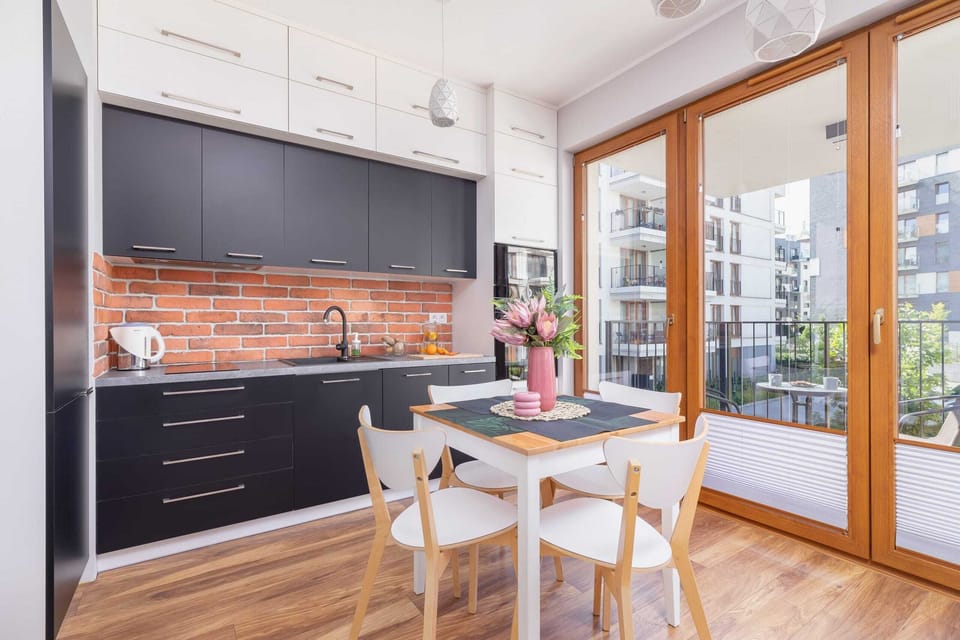A dining area with a wooden table and chairs, an exposed brick wall, and a modern kitchen with sleek black cabinetry.