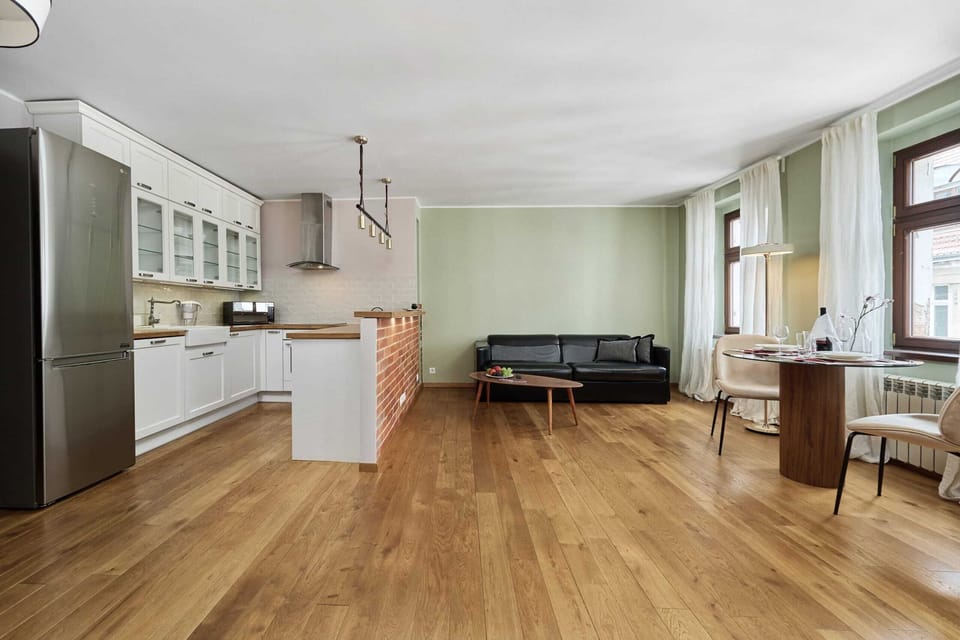 Spacious living room with a wooden floor, light curtains, and a black sofa. The room is bright and filled with natural light.