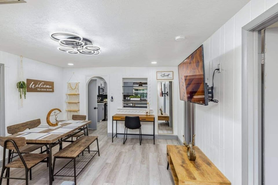 The living room is a modern and cozy space that combines a dining area and a relaxing zone. It features a wooden table with chairs and a bench, a desk for working or studying, a sofa bed, a TV, and stands out for its excellent lighting