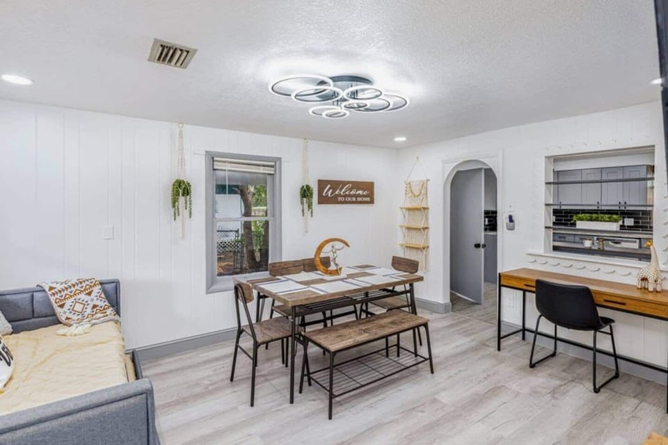 The living room is a modern and cozy space that combines a dining area and a relaxing zone. It features a wooden table with chairs and a bench, a desk for working or studying, a sofa bed, a TV, and stands out for its excellent lighting
