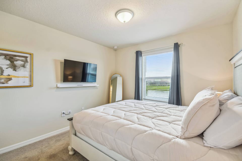Natural light floods this cozy bedroom, highlighting the soft, neutral tones and plush bedding. Enjoy the convenience of a mounted TV for entertainment, while large windows offer a view that connects you to the outside world.