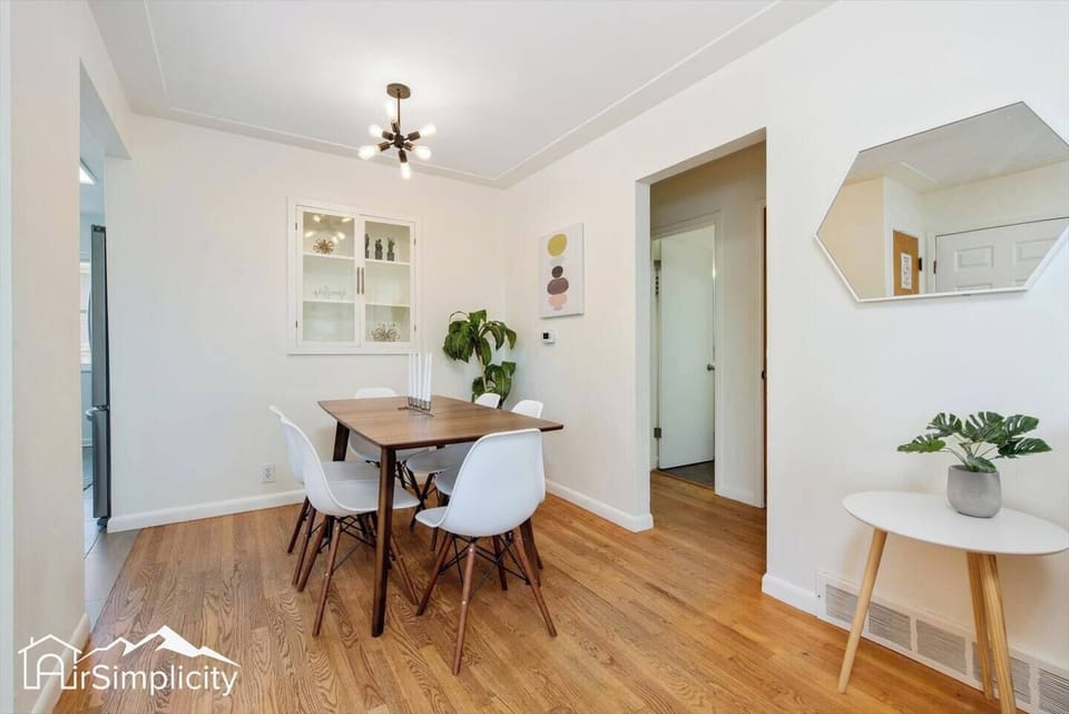 - Clean lines and mid century details define this warm dining setting
- Wall art, hanging light, and greenery bring a bright, curated feel
- Hardwood floors and white walls create an airy space for group meals