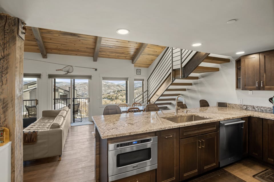 A modern kitchen with granite countertops, stainless steel appliances, and an open layout leading to a living area with a vaulted wooden ceiling and large windows offering a mountain view.