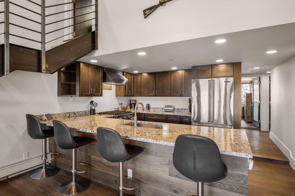 Modern kitchen with granite countertops, stainless steel appliances, and four black bar stools. A staircase with a metal railing leads to an upper level. The room is well-lit with recessed lighting.