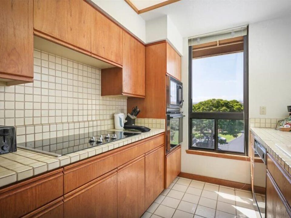 Your fully-equipped kitchen features warm wood cabinetry and tiled countertops, with a window framing lush greenery—perfect for preparing meals while soaking in natural light.