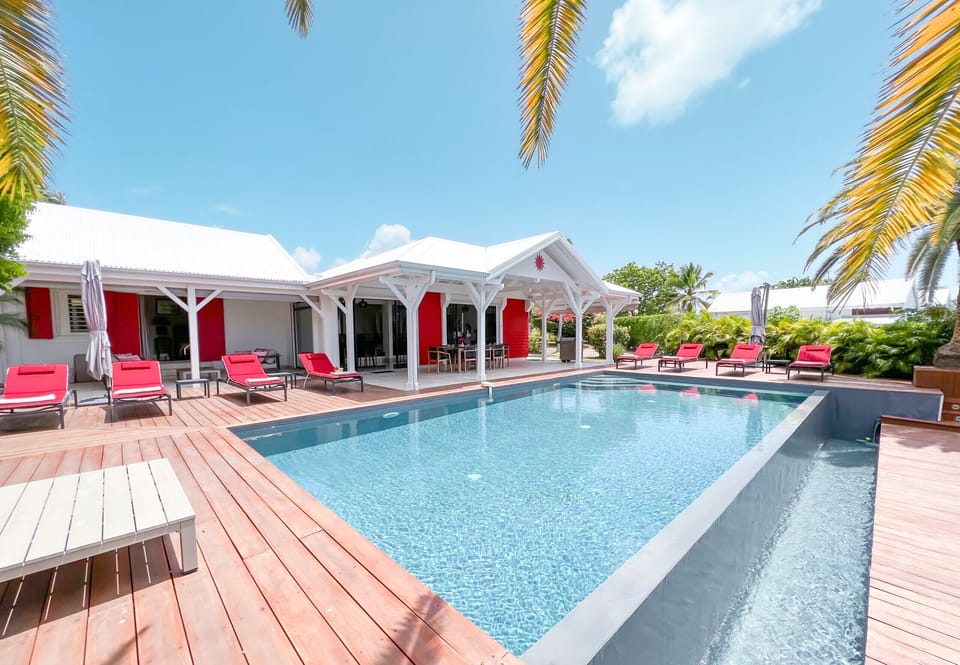 Discover our luxury villa in the West Indies, offering a captivating view of the infinity pool surrounded by red deckchairs. Admire the elegant façade of the house, a symbol of the refinement and comfort of this exceptional residence. Immerse yourself in absolute luxury, where relaxation meets sophistication. Book now for an unforgettable experience.