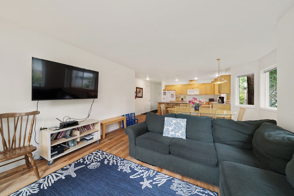 Relax in your open-concept living space with a cozy sofa, flat-screen TV, and fully-equipped kitchen. Sunlight fills the room through windows framing peaceful woodland views.