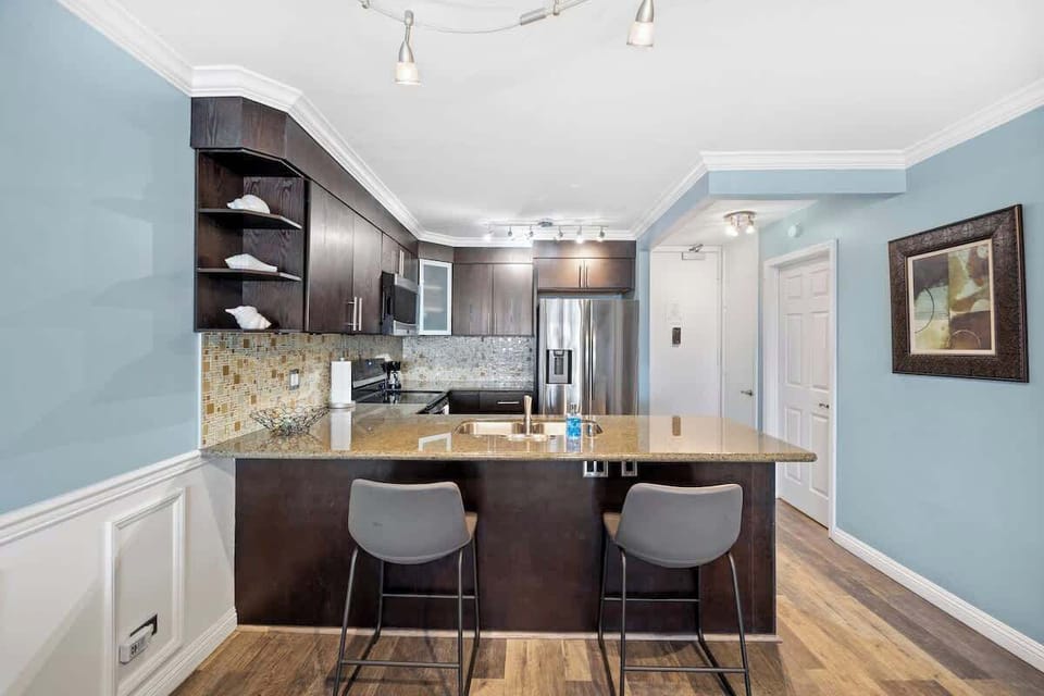 Modern kitchen with dark cabinets, stainless steel appliances, granite island with bar seating, and mosaic backsplash. 