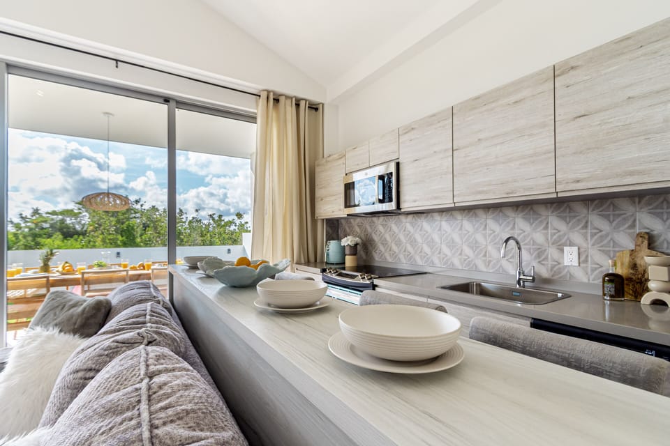 Picture yourself cooking in this bright, airy kitchen where modern design meets functionality. Light wood cabinetry, stylish tile backsplash, and expansive windows frame lush tropical views, making meal prep feel like a serene retreat.