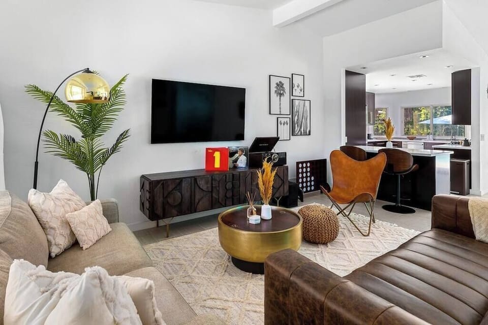 Bright living room with wall-mounted TV, record player console, leather butterfly chair, and open sightline to kitchen — laid-back Palm Springs cool.