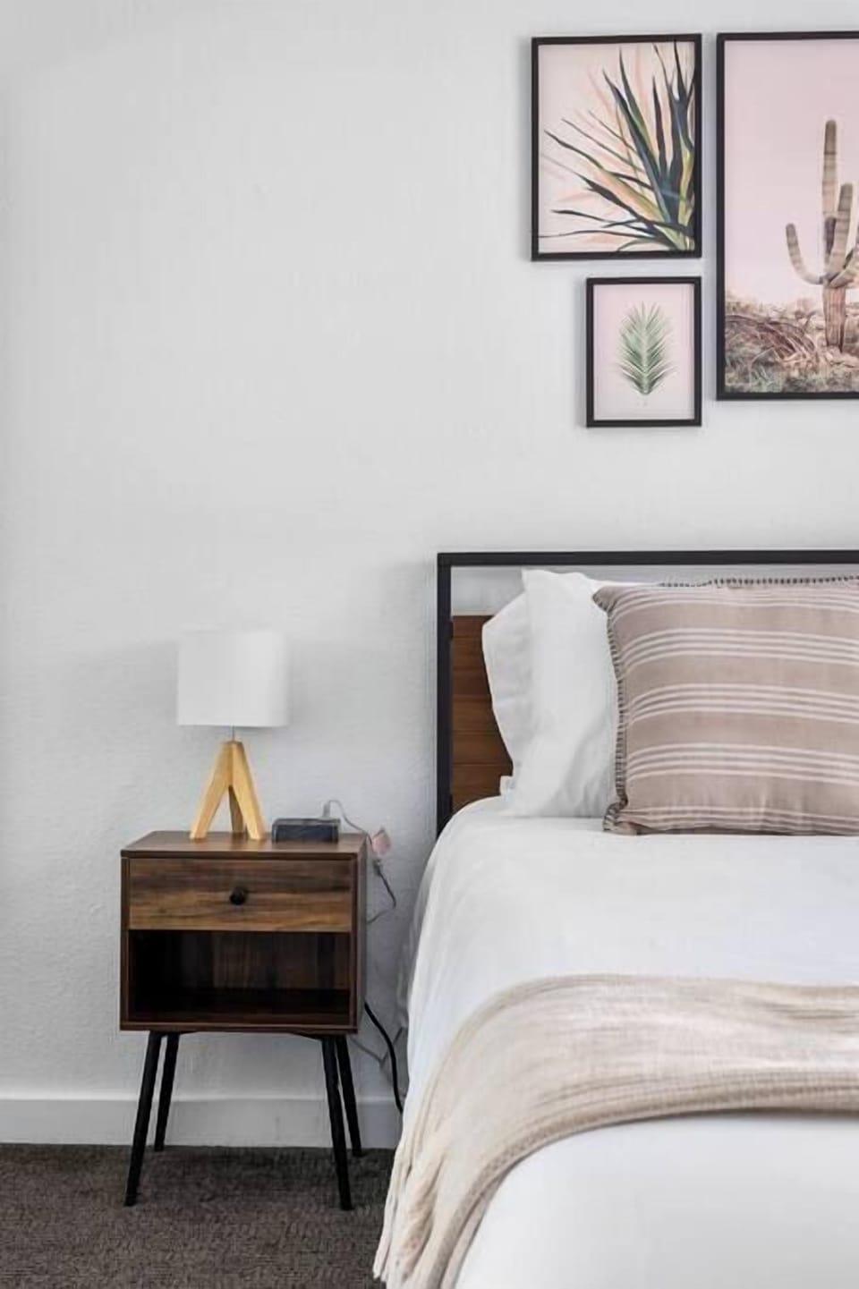 Bedroom detail — wood nightstand, desert botanical gallery wall, and crisp white linens with woven throw. Relaxed Palm Springs mood nailed.