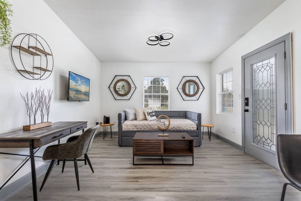 Living room: 
A modern and bright living room featuring a sleek daybed with decorative pillows as a focal point. The space is tastefully furnished with a stylish wooden desk and chair, making it a perfect spot for work or study. The room has light-colored flooring and a glass-paneled door, allowing natural light to fill the area. Unique wall art and geometric mirrors add a touc...