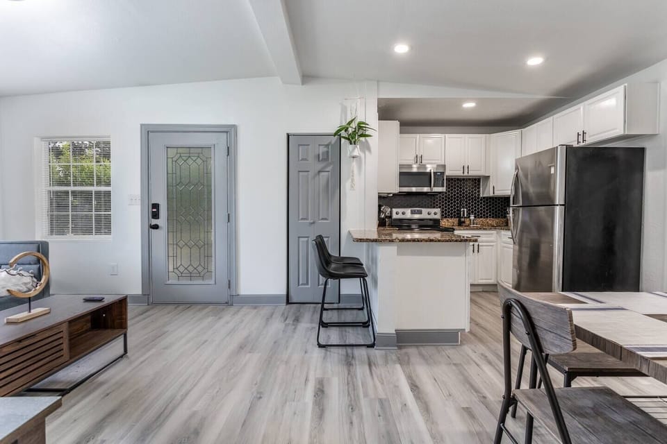 A modern, open-concept kitchen and living area featuring light wood flooring that adds warmth and brightness to the space. The kitchen boasts white cabinetry, stainless steel appliances, and a sleek black backsplash for a contemporary look. A cozy breakfast bar with a high stool provides a casual dining spot, while a hanging plant introduces a touch of greenery. The glass-panel...