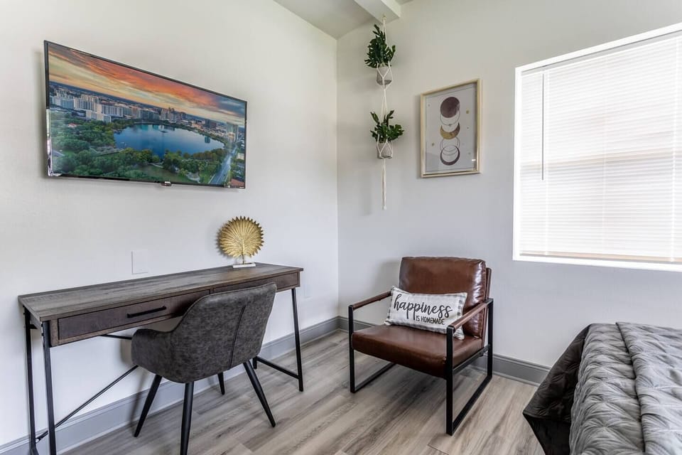 Bedroom 2: A stylish and cozy corner of the bedroom, featuring a sleek wooden desk paired with a comfortable chair, creating an ideal workspace. Above the desk, a flat-screen TV displays a serene cityscape, adding both functionality and entertainment. The space is further enhanced by a brown leather armchair adorned with a decorative pillow that reads "happiness." Natural light...
