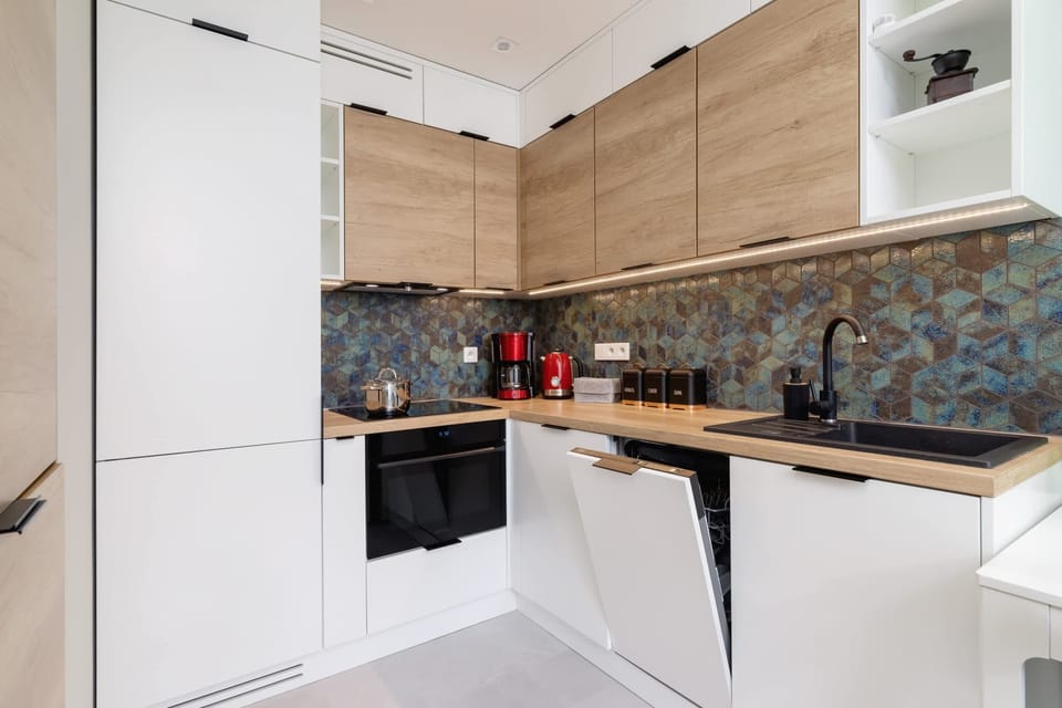 A modern kitchen with light wooden fronts, white cabinets, and a decorative stone-patterned backsplash, fully equipped with appliances.
