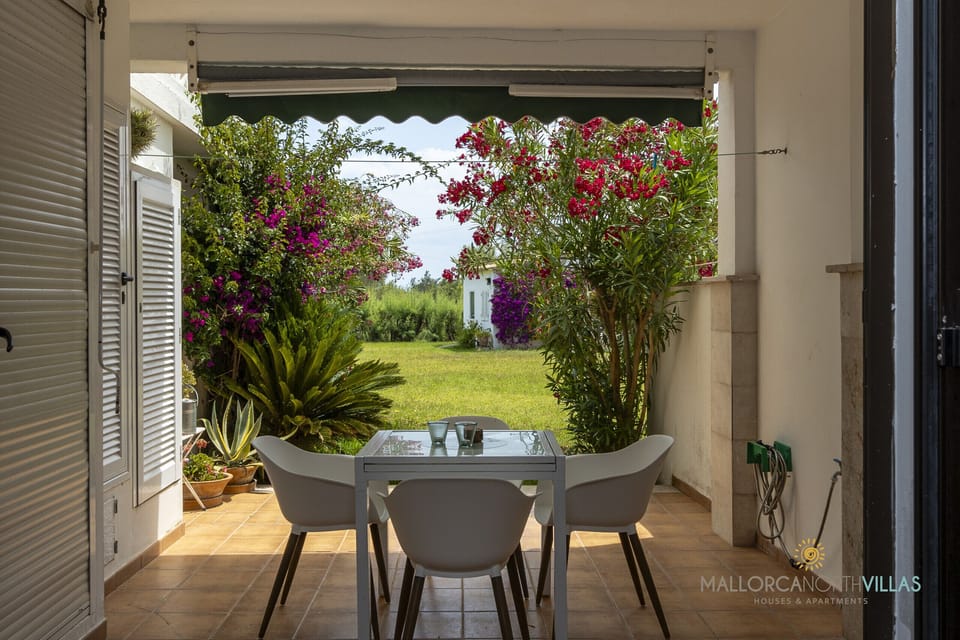 Covered terrace with outdoor dining table and views of a spacious garden with Mediterranean flowers and plants.