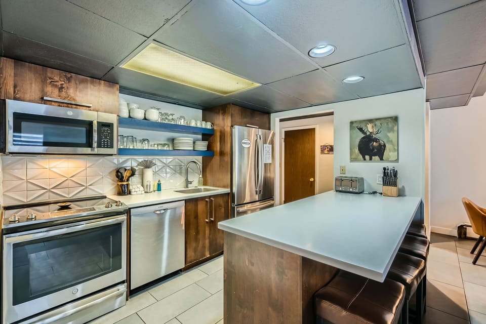 Modern kitchen with stainless steel appliances, white countertops, wooden cabinets, and a central island with bar stools. A painting of a moose is on the wall adjacent to the refrigerator.