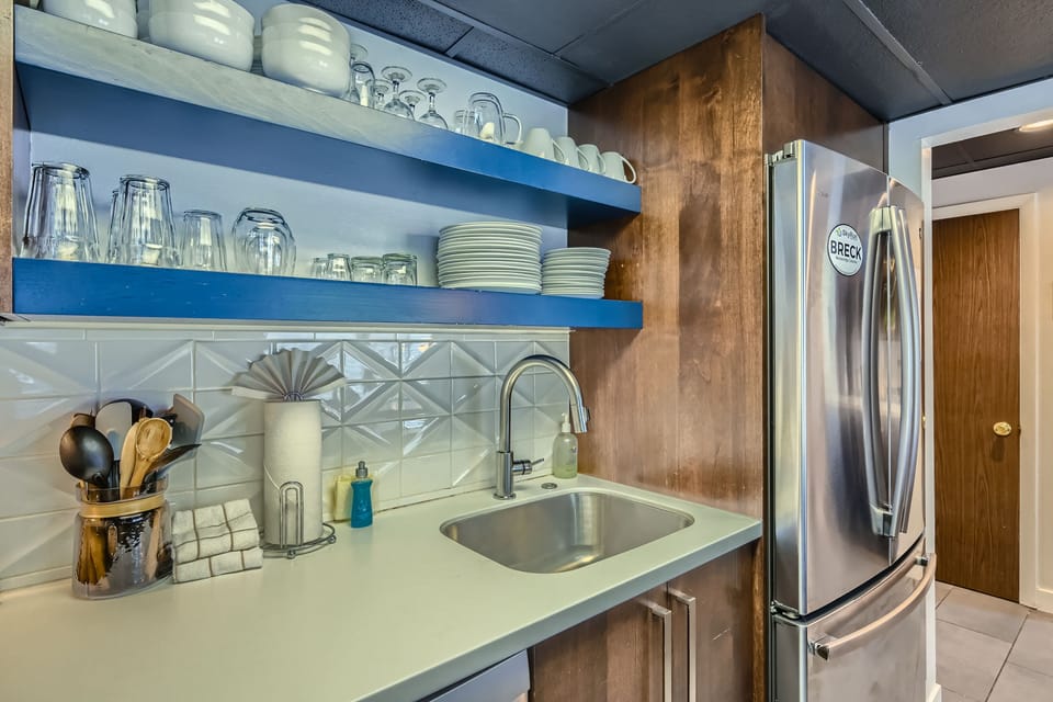 A kitchen area with open shelving holding dishes and glasses, a stainless steel refrigerator, a sink, utensils in a holder, a soap dispenser, and a tiled backsplash.