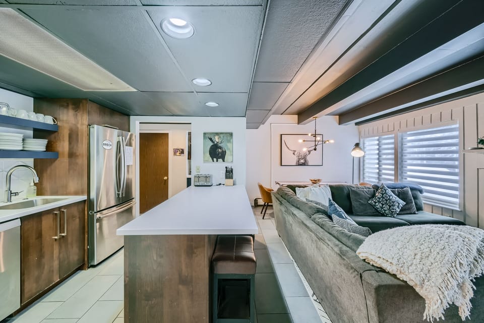 A modern kitchen and living room with a wooden island, stainless steel refrigerator, shelves with dishes, gray sofa, cushioned chairs, wall art, and large windows with blinds.