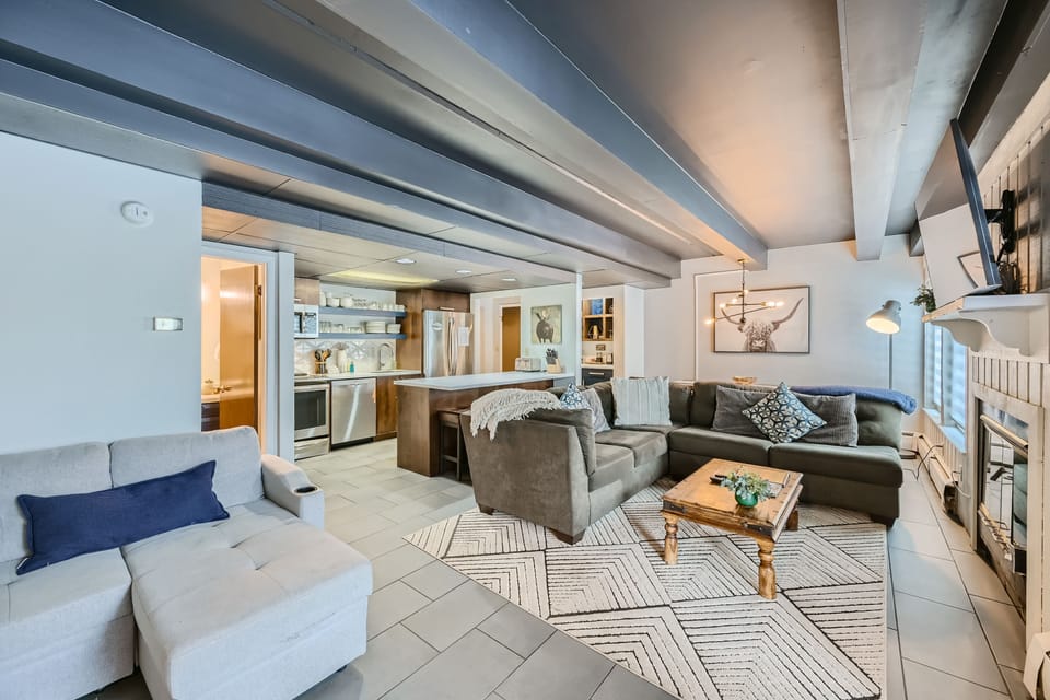 A modern living space featuring a sectional sofa, coffee table, and patterned rug. The room has exposed beams, an open kitchen with bar seating, and a wall-mounted TV above a fireplace.