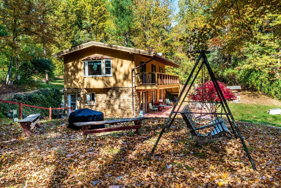 Backyard with metal-framed wooden swing, picnic benches, charcoal grill, and stone-and-wood cabin exterior.