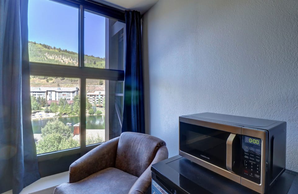 A room with a large window showcasing a hillside view, a brown armchair, and a microwave oven positioned on top of a mini-fridge.