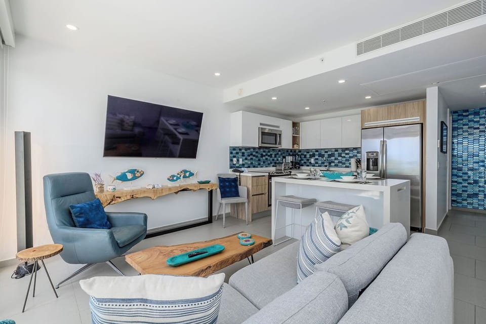 Relax in your light-filled open-plan living area with plush coastal-inspired seating, stylish blue accents, and a fully equipped modern kitchen ready for your culinary adventures.