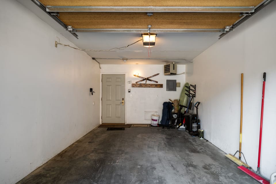 A empty garage with a concrete floor, white walls, and an open garage door. Various tools and cleaning supplies are stored along the right wall. A closed door is seen at the far end of the garage.