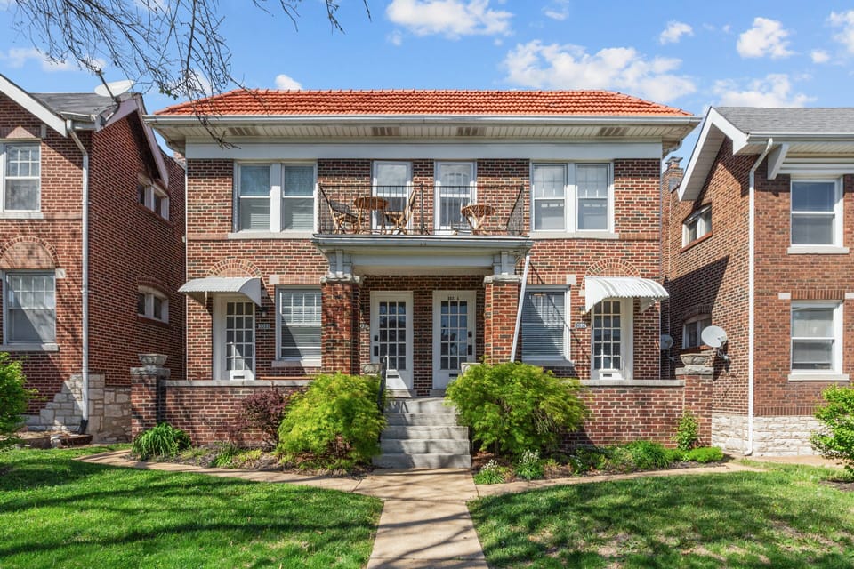 The daytime exterior shows a two-story brick residential building with central concrete steps, covered porch, upper balcony, striped entry awnings, landscaped beds, front lawn, and neighboring homes.