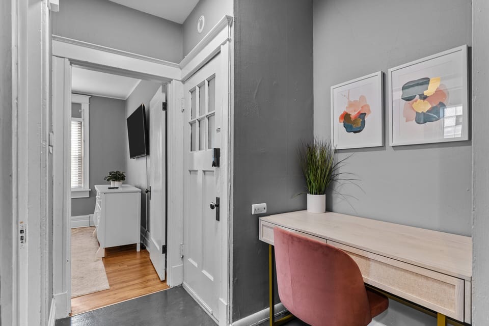A dedicated light-wood desk with pink upholstered chair sits in the hall beneath two framed prints, with a tall potted plant and bedroom doorway nearby.