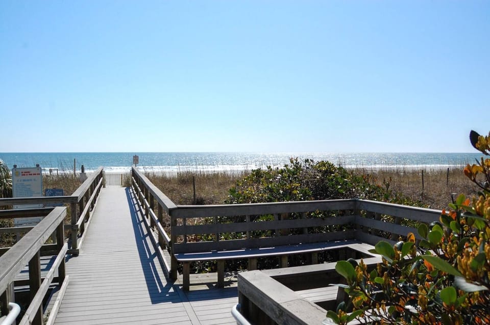 A wooden boardwalk leads directly to pristine sandy shores and sparkling ocean waters under clear blue skies.