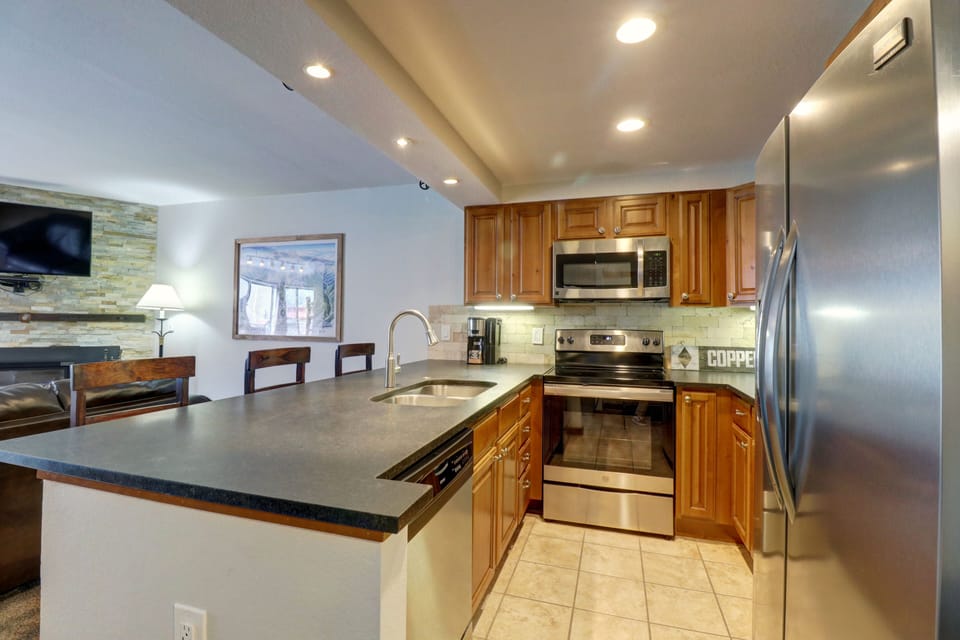 Modern kitchen with stainless steel appliances, wood cabinetry, granite countertops, a double sink, and tiled flooring. An open view of the living room area with a wall-mounted TV is visible in the background.