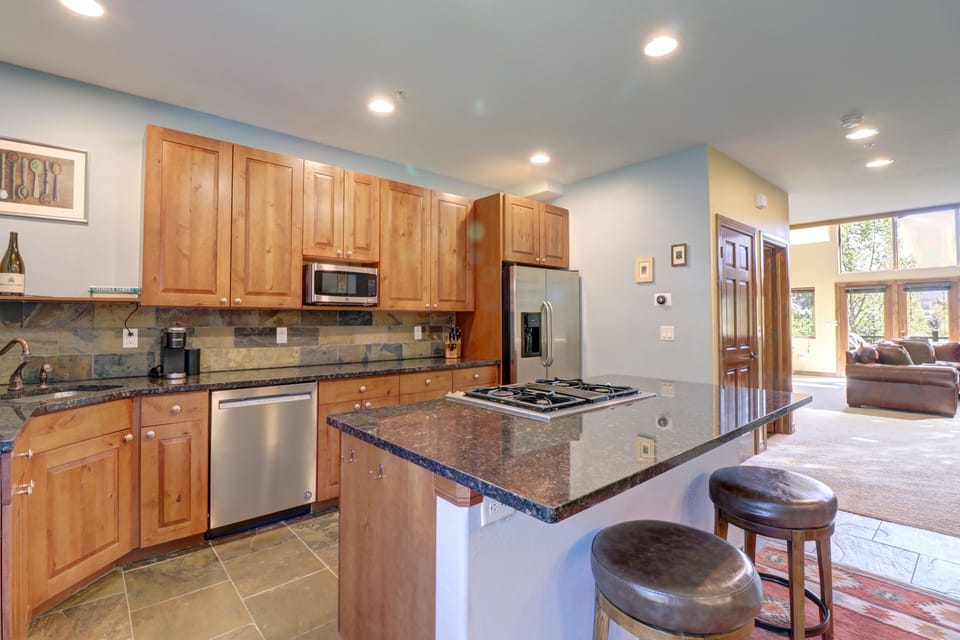 Modern kitchen with wooden cabinets, stainless steel appliances, a black granite countertop, and an island with two bar stools. Living room visible in the background. Ample natural light throughout.