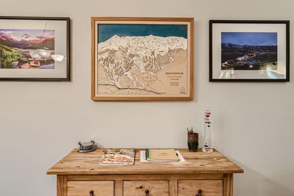 A wooden cabinet with a few magazines and small objects sits below a framed map of Breckenridge and two framed photographs of mountainous landscapes on a beige wall.