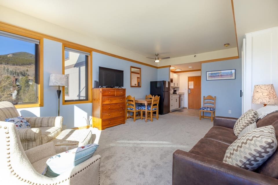 A brightly lit living area features a couch, armchair, wooden dresser, dining table with chairs, and wall-mounted TV. The adjacent kitchenette includes a refrigerator and cabinetry.