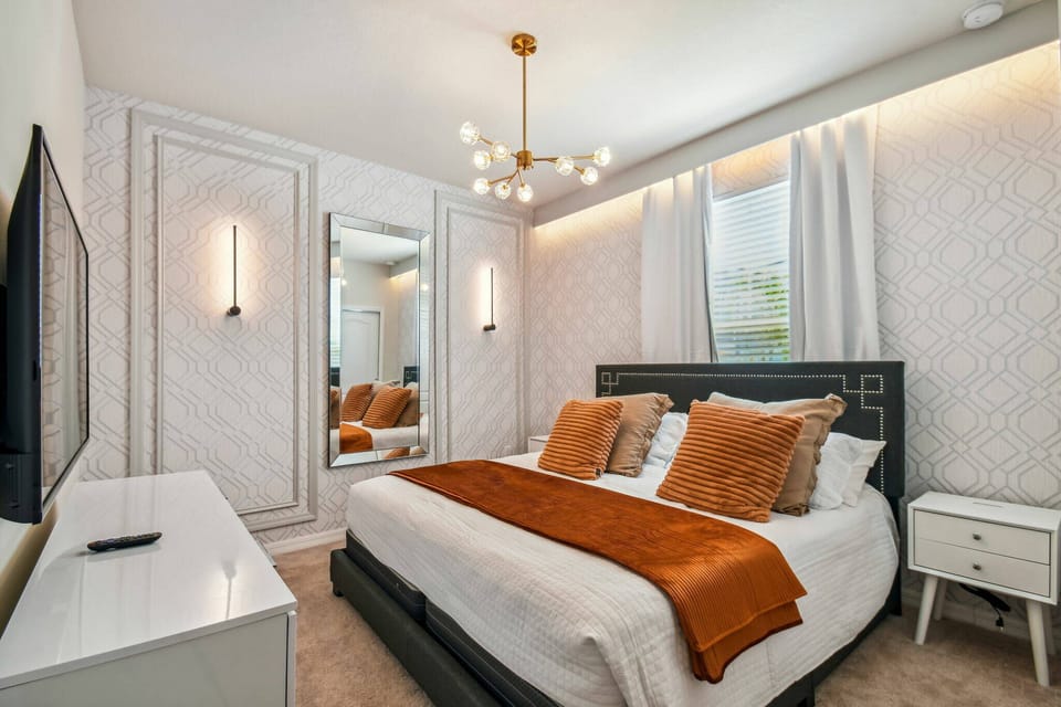 Experience a beautifully designed bedroom featuring a plush king-sized bed with rich orange accents and soft white linens, complemented by a modern chandelier and sleek mirrored wardrobe, all set in a warm, inviting ambiance.